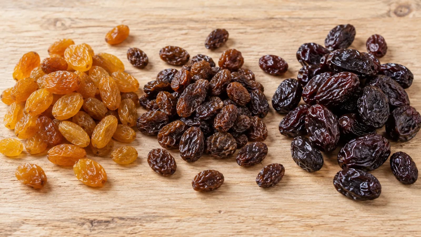 Tres montones de pasas de uva sin semilla sobre una tabla de madera clara: Sultanina dorada, Thompson Seedless marrón oscura y Flame Seedless rojiza casi negra, de mayor calibre